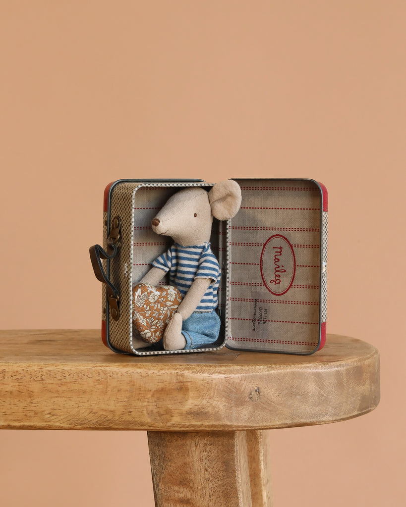 A Maileg LouiMax On the Go Suitcase Set, featuring a mouse in a striped shirt and blue pants inside an open metal suitcase, is displayed on a wooden bench with a plain beige background.