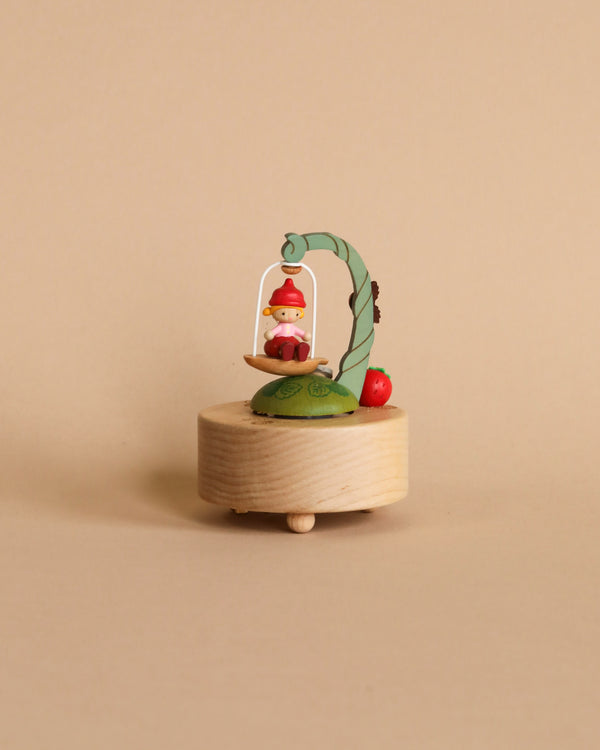 A small, whimsical Mini Fairy On Swing hand-cranked music box featuring a tiny gnome sitting under a leafy arch, with decorative apples beside him, all mounted on a round wooden base.