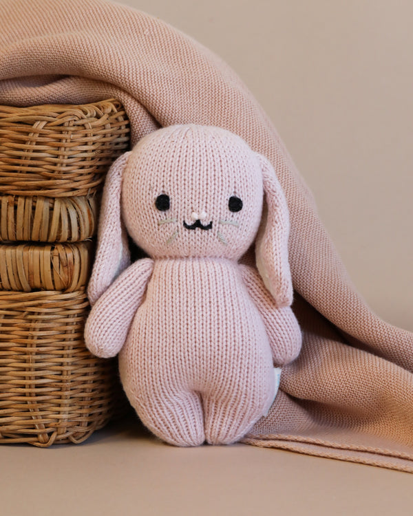 A pink hand-knit Cuddle + Kind Baby Bunny stuffed toy sitting in front of a woven basket, partially covered by a soft beige blanket. The bunny features cute eyes and a smiling mouth stitched in black.