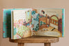 The Birthday Music Book is open on a wooden stool, showing illustrated pages of a cozy bedroom with two beds, colorful bedding, toys, and a pajama-clad mouse. Musical notes and text—like musical clips—appear on the left page.