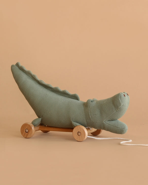 The Pull-Along Croc is a soft, green, crocodile-shaped toy with wooden wheels and a white string—perfect for developing gross motor skills. From Egmont Toys, it’s shown on a plain beige background.