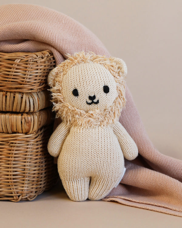 A cute Cuddle + Kind Baby Lion toy hand-knit from Peruvian cotton yarn stands in front of a woven basket partially covered by a soft pink blanket against a beige background.