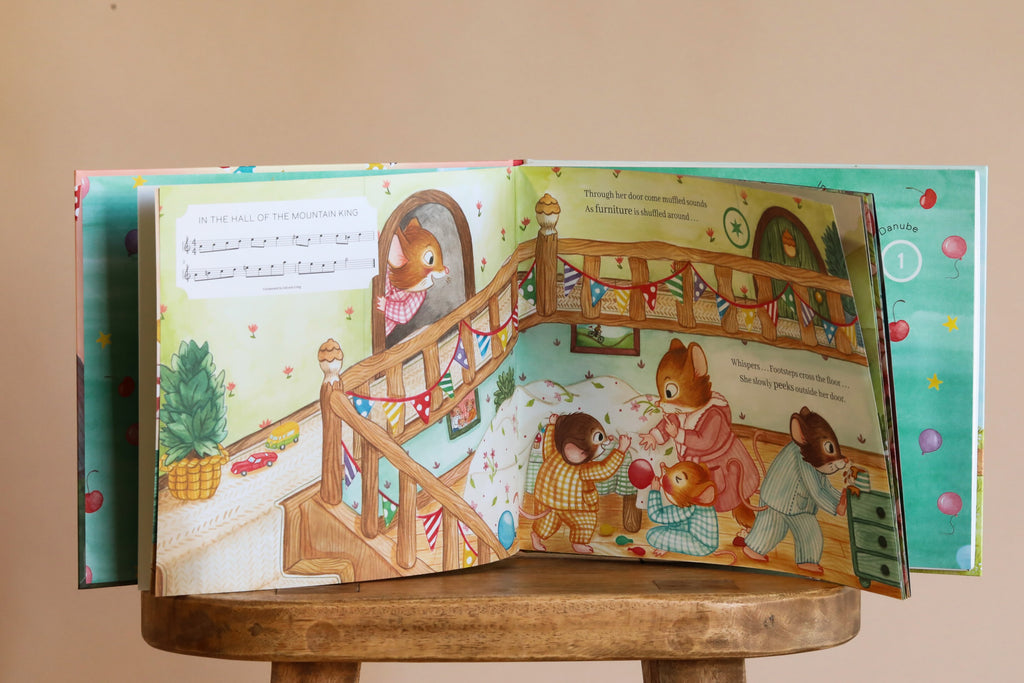 The Birthday Music Book is open on a wooden stool, displaying illustrated animals celebrating in a colorful, festive home. Both pages feature party scenes and sheet music inspired by classical masterpieces.