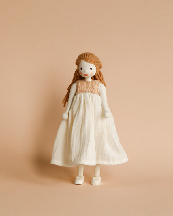 A Ferne Wooden Doll with red hair and a wide smile, wearing a long white dress, stands against a soft beige background.