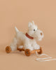 The Pull-Along Lola Dog is a plush white toy with curly fur, a red plaid collar, and wooden wheels. It features a white pull string, perfect for toddlers to develop gross motor skills. Shown here on a beige background.