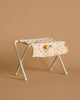 The Maileg Nursery Table, Baby Mouse - Off White is a compact, foldable stool featuring a metal frame and covered with beige fabric adorned with colorful dots. The fabric includes two front pockets, housing a tiny doll and a small toy bottle. It has the appearance of a nursery table, seamlessly blending into its smooth tan backdrop.