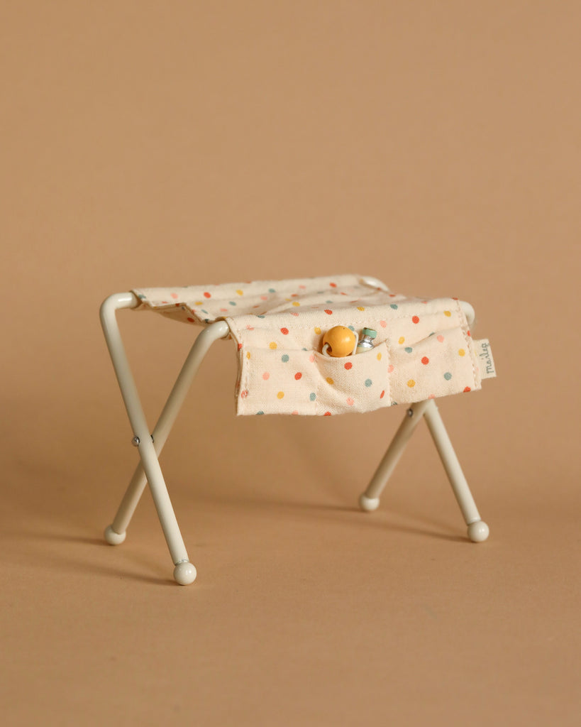 The Maileg Nursery Table, Baby Mouse - Off White is a compact, foldable stool featuring a metal frame and covered with beige fabric adorned with colorful dots. The fabric includes two front pockets, housing a tiny doll and a small toy bottle. It has the appearance of a nursery table, seamlessly blending into its smooth tan backdrop.