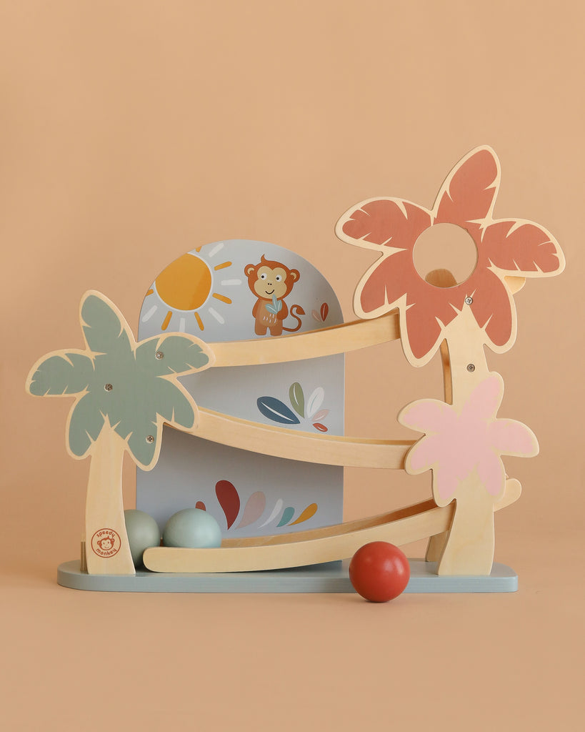 The Wooden Ramp Racer is made from FSC certified wood, features three pastel palm trees, a monkey illustration, and four colored balls (green, blue, gray, red), designed for fine motor skill development against a peach background.