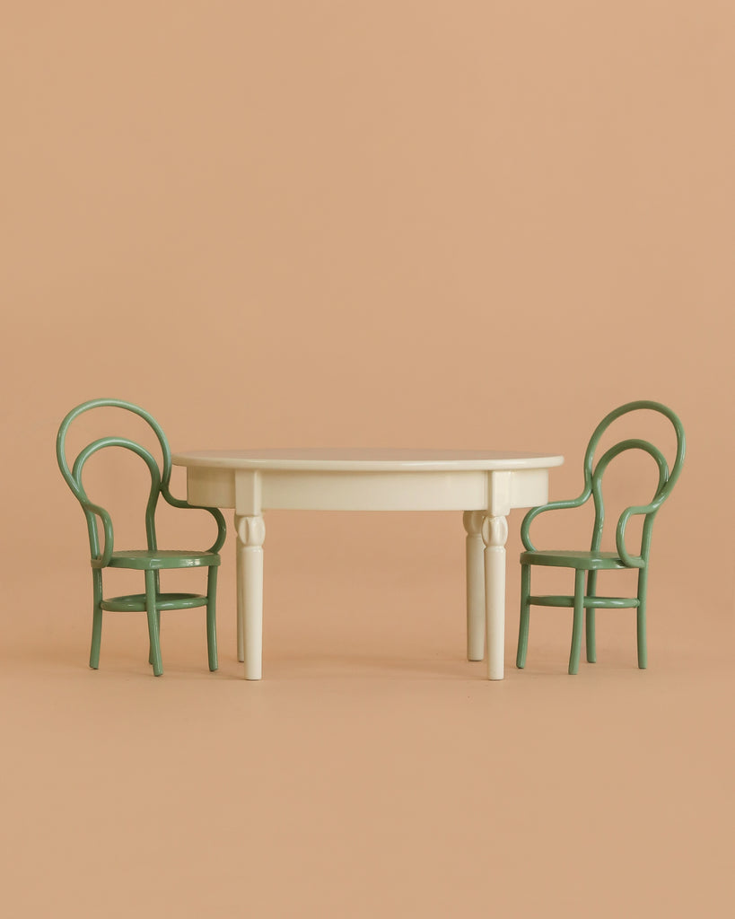 A Maileg Dining Set in cream, sized for mice, includes a vintage table placed between two green round-backed chairs against a plain beige background.