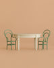 A minimalist scene featuring the Maileg Little Brother and Sister Dining Set, with a cream round vintage table and two green curved-back chairs arranged on either side, set against a solid beige background.