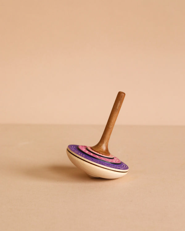 The Mader Bonbon Spinning Top - Purple showcases a purple and pink pattern, elegantly balanced on its point against a beige background—an embossed, classic wooden toy inspired by Austrian craftsmanship.