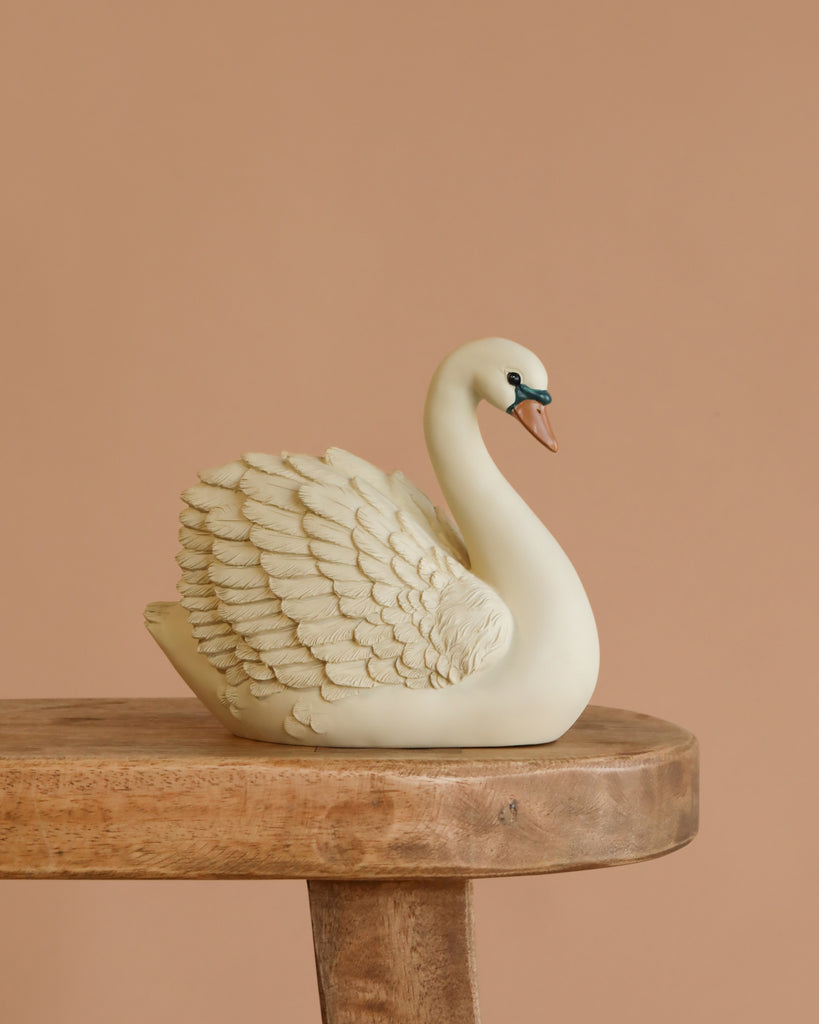 The Konges Sloejd Money Bank - Swan, a beige ceramic piggy bank with detailed feathers, sits on a wooden surface against a plain light brown background, offering a charming and stylish way to save money.