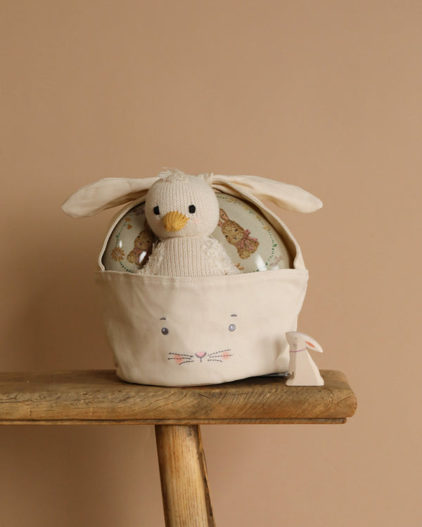 A soft crocheted duck toy rests inside the Easter Basket, featuring bunny ears and a face, placed on a wooden stool. Next to it is an heirloom-quality wooden Easter bunny cutout, all set against a warm beige wall that enhances the charm of the scene.