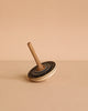 The Mader Bonbon Spinning Top - Black, with its embossed pattern, rests tilted on a beige surface against a plain beige background, capturing the timeless charm of classic Austrian toys.