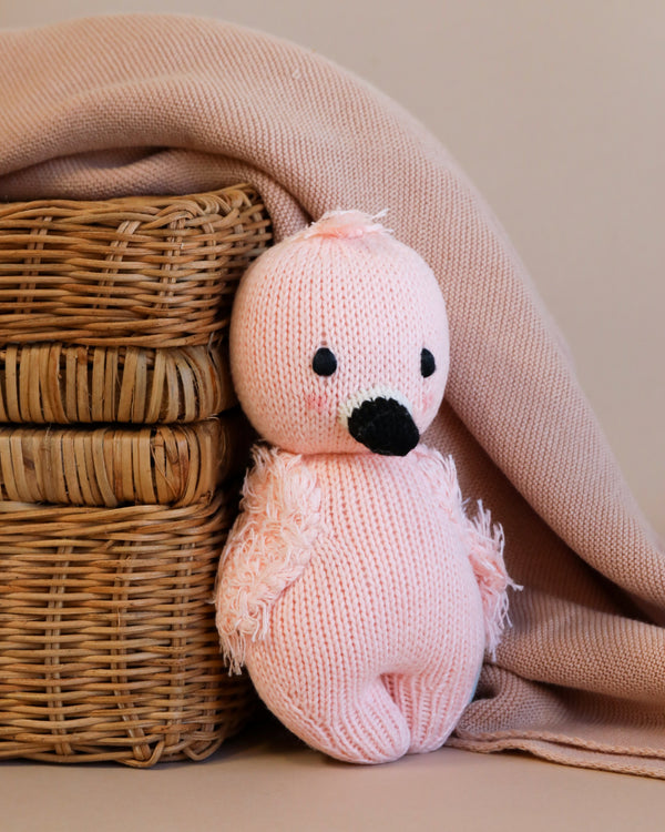 A handmade pink knitted Cuddle + Kind Baby Flamingo plush toy sits in front of a wicker basket, partially covered by a soft beige cloth. Made from Peruvian cotton yarn, the toy features a darker pink