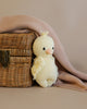 A soft, hand-knit toy baby duckling leaning against a small wicker basket, partially covered by a textured pink blanket on a neutral background.