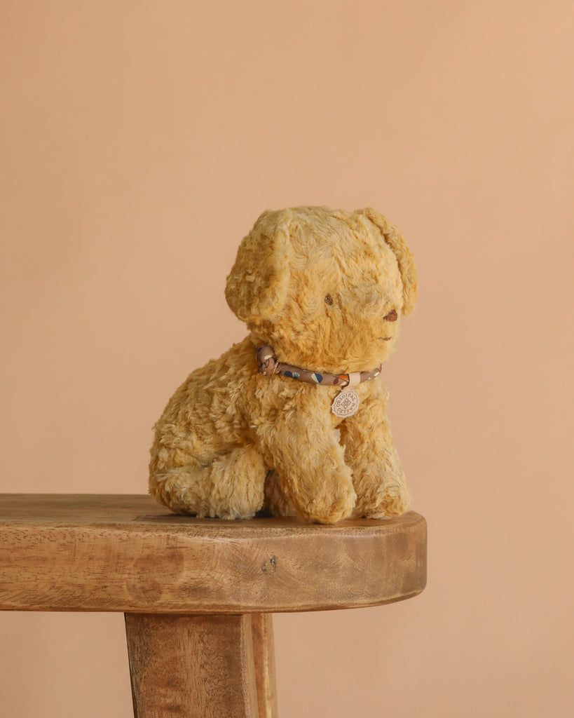 The Maileg Puppy, Small - Soft Sand, a vintage-style stuffed dog with a beaded collar, sits on the edge of a wooden bench against a plain beige background.