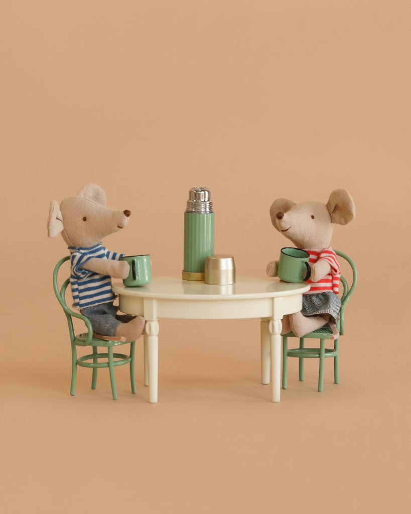 The Maileg Little Brother and Sister Dining Set features two mice in striped shirts sitting on green chairs at a vintage dining table with green mugs, a miniature thermos, and cup, all against a beige background.