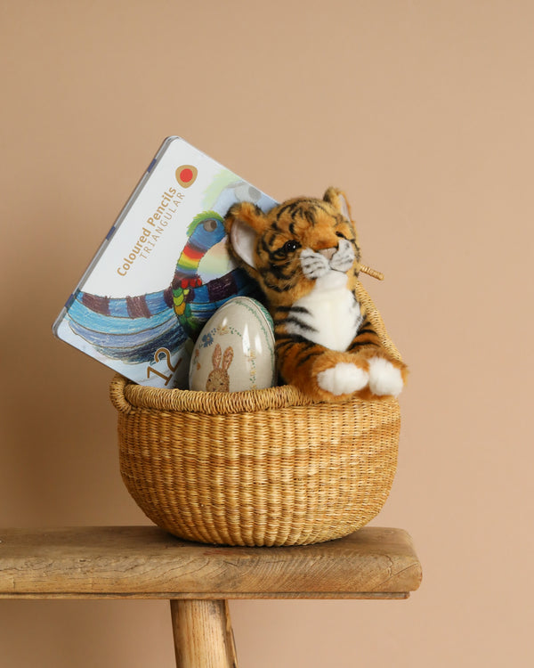 The Handmade Easter Basket sits on a wooden stool against a beige background, filled with a children's book featuring a parrot on the cover, a Tiger Cub Stuffed Animal toy, and a decorated egg.