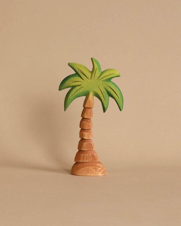 The Bumbu Wooden Small Palm Tree features a charming, stacked brown trunk and vibrant green fronds on top, all set against a light beige background. Its simple and minimalist design lends the figurine a playful and artistic appearance, making it perfect for imaginative play or as a handcrafted wooden toy.