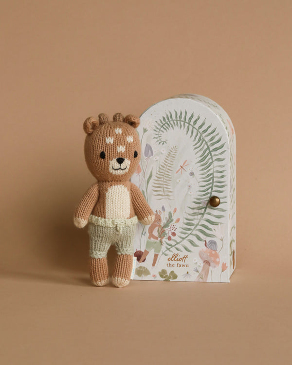 A Cuddle + Kind Tiny Elliott The Fawn standing next to a small illustrated door decorated with floral and fauna motifs on a beige background, crafted using sustainable fair trade practices.
