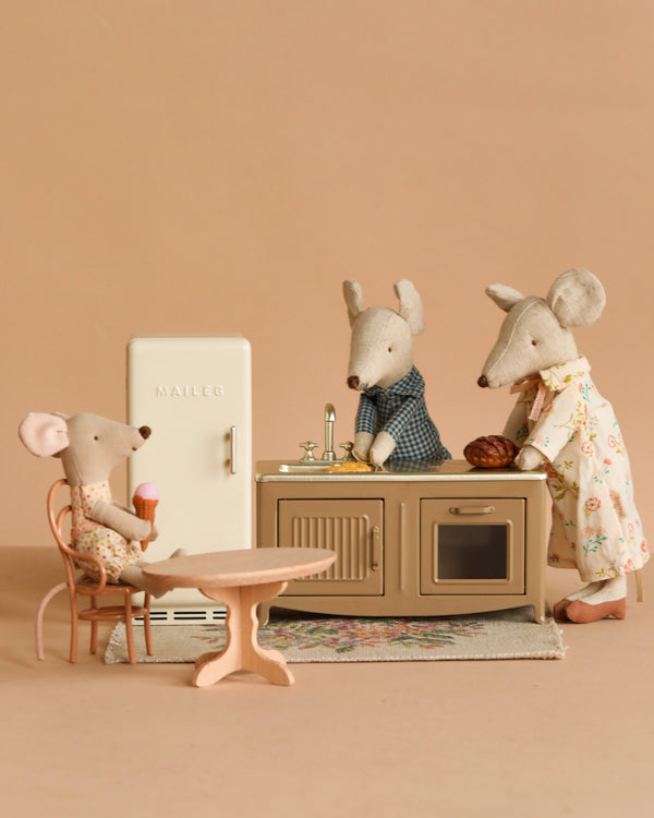 Three Maileg mice from the Afternoon with Grandparents Set are in a mouse house kitchen, with one eating ice cream at the table and two by the sink—one in an apron, one in a robe—amid charming Maileg kitchen accessories.