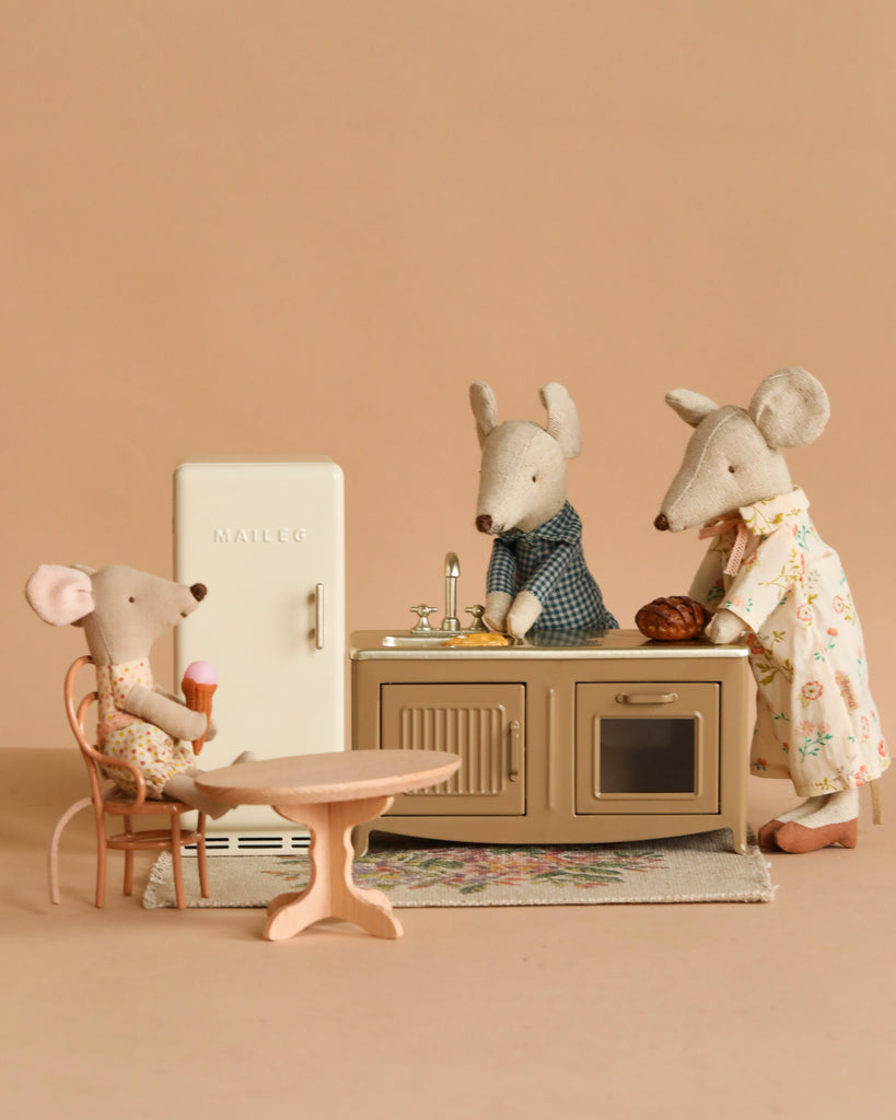 Three Maileg mice from the Afternoon with Grandparents Set are in a mouse house kitchen, with one eating ice cream at the table and two by the sink—one in an apron, one in a robe—amid charming Maileg kitchen accessories.