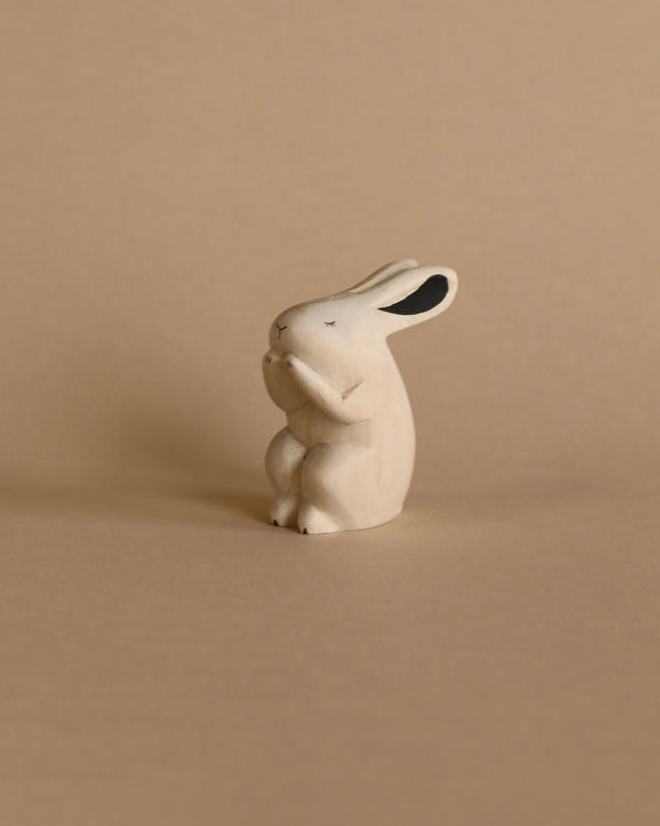 A small, white Handmade Tiny Wooden Forest Animal - Bunny with black accents on its ears and eyes, positioned against a neutral beige background.