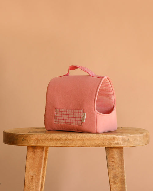 The Maileg Pet Carrier - Coral, a small pink cotton-linen carrier with a grid-patterned pocket and handle, sits on a wooden stool against a plain beige background.