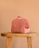 The Maileg Puppy in Coral Pet Carrier - Dusty Powder, a small pink fabric pet carrier for kids with a handle and mesh window, is displayed on a wooden stool against a plain beige background.