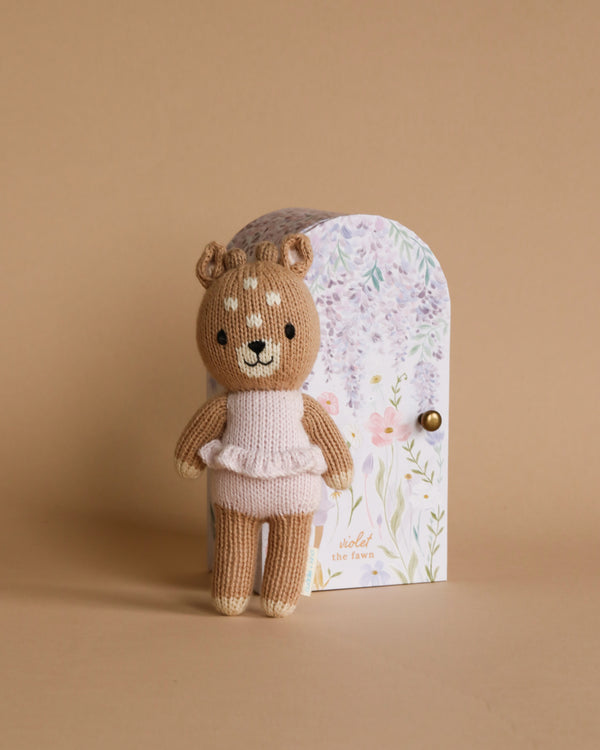 A small hand-knit Cuddle + Kind Tiny Violet The Fawn wearing a white sweater stands in front of a floral-printed birthday card with a rounded top, set against a soft beige background.