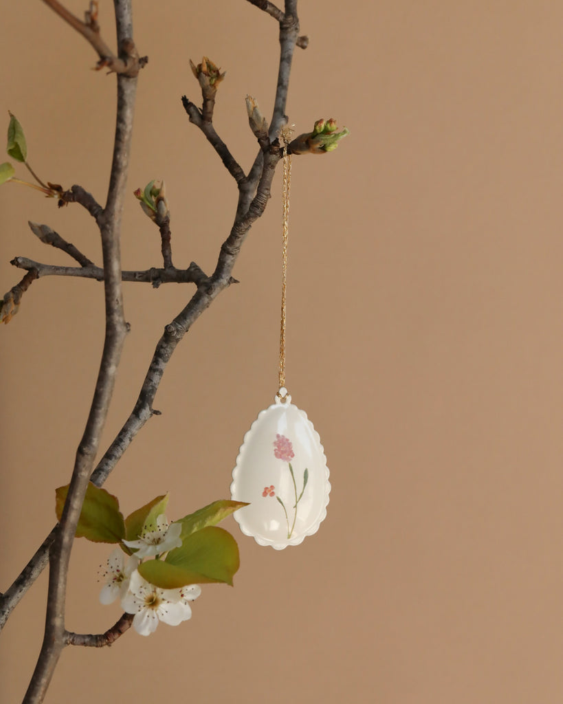 The Maileg Metal Ornament - Easter Egg, Rose is a hand-painted white egg with a pink flower design, hanging from a gold chain on a budding tree branch with green leaves and white blossoms, set against a plain beige background.