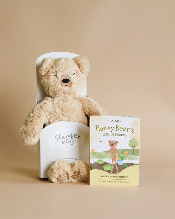 A plush teddy bear sits snugly in a white tube labeled "Slumberkins Honey Bear Kin," adjacent to a children's book titled "Honey Bear's Gifts of Nature - A Lesson in Cultivate Gratitude.