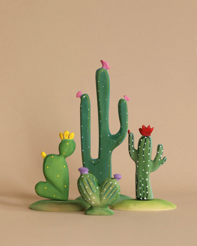 The Bumbu Wooden Cacti Set features a delightful array of clay and wooden cactus sculptures, showcasing different shapes and vibrant colors. One cactus has yellow dots with yellow flowers, another green one adorns small pink birds, a third is speckled with white spots and topped with a red flower, and the last is a short, wide piece crowned with purple flowers.