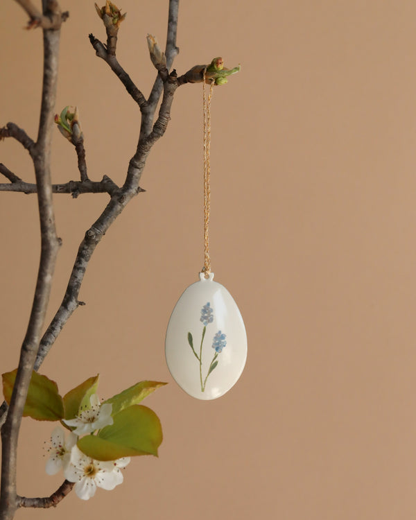 The Maileg Metal Ornament - Easter Egg, Blue features a decorative hand-painted blue floral design and hangs from a gold chain on a tree branch with white blossoms, set against a beige background.