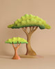 Two stylized wooden trees with green canopies and brown trunks stand on a beige surface—perfect for imaginative play. This handcrafted set pairs beautifully with the Bumbu Handmade Wooden Prehistoric Set.