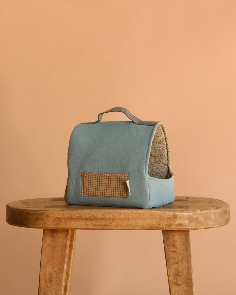 The Maileg Pet Carrier - Dusty Blue, a small cotton linen carrier with handle, sits on a wooden stool against a peach background. It features a soft, patterned interior and side pocket—ideal for carrying stylish pet accessories.