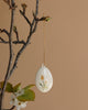 A decorative hand-painted ceramic egg with floral designs hangs by a gold string among green leaves and white blossoms on a bare branch, set against a beige background—ideal for pairing with the Maileg Metal Ornament - Easter Egg, Yellow.