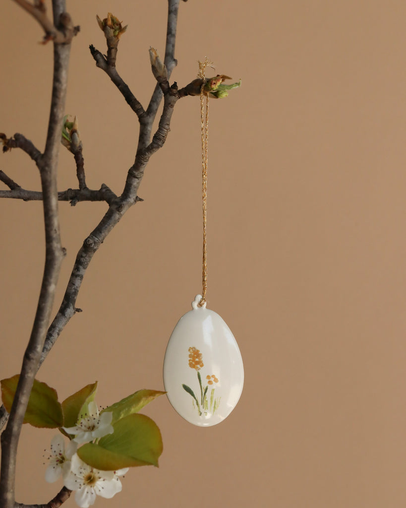 A decorative hand-painted ceramic egg with floral designs hangs by a gold string among green leaves and white blossoms on a bare branch, set against a beige background—ideal for pairing with the Maileg Metal Ornament - Easter Egg, Yellow.