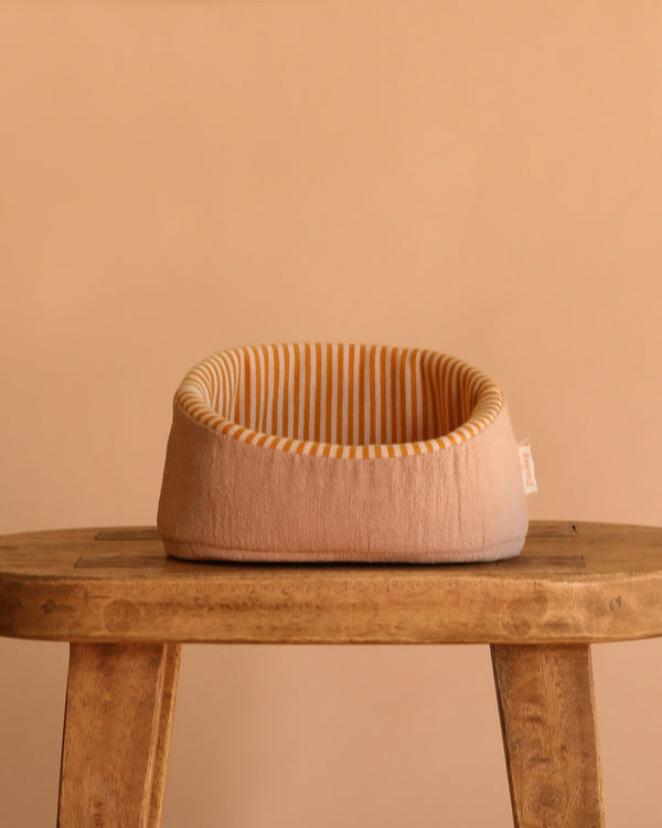 The Maileg Pet Basket - Powder, a small round beige bed with yellow and white stripes inside, sits on a rustic wooden stool against a plain beige background—perfect for Maileg puppies.