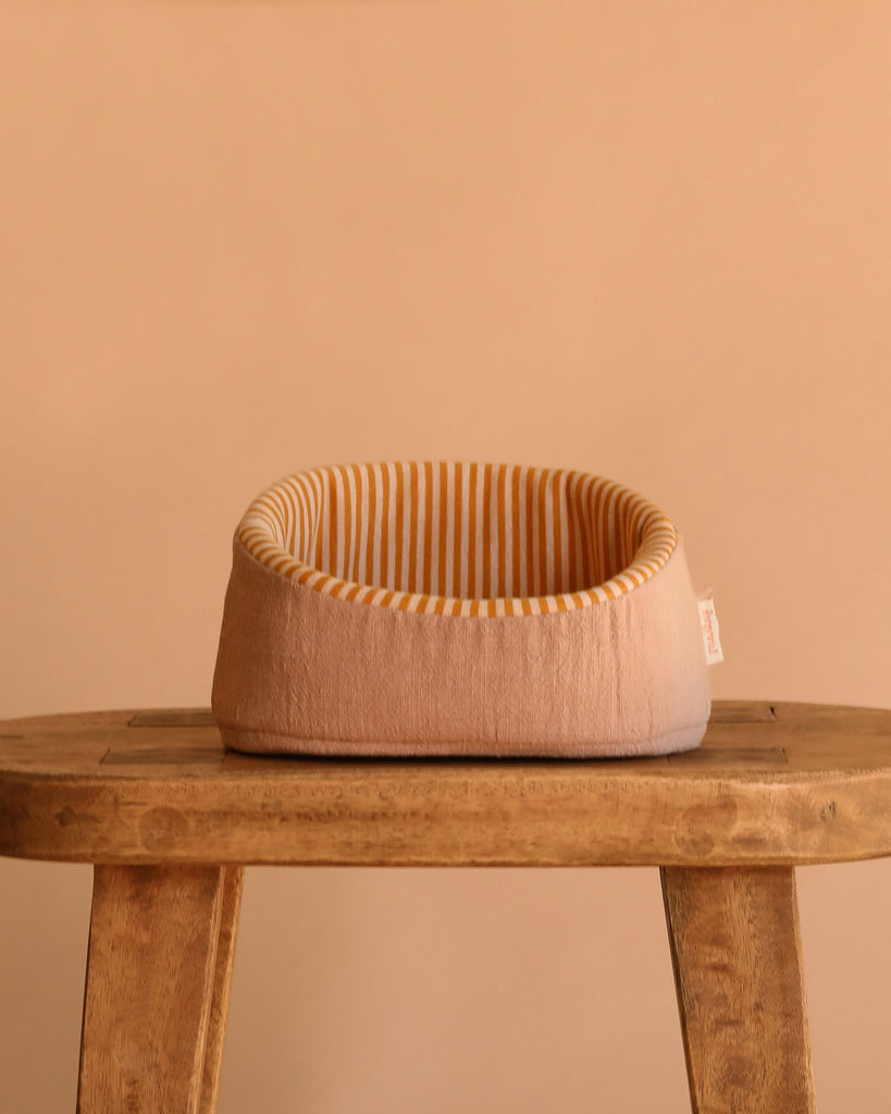 The Maileg Pet Basket - Powder, a small round beige bed with yellow and white stripes inside, sits on a rustic wooden stool against a plain beige background—perfect for Maileg puppies.