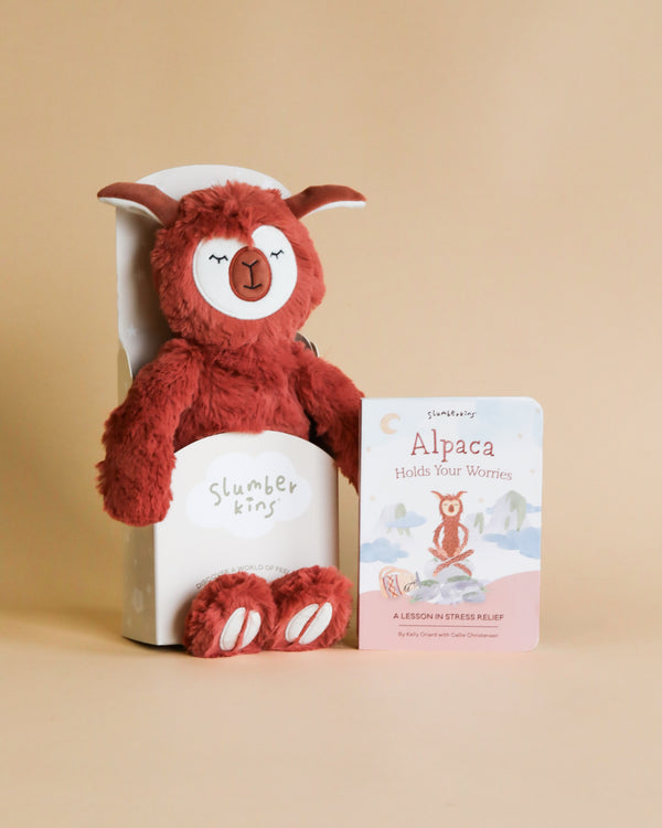 A Slumberkins Alpaca Kin + Lesson Book on stress relief designed for anxiety management, in a sitting position, placed beside a small card titled "alpaca holds your worries" with a backdrop of a light cream wall.