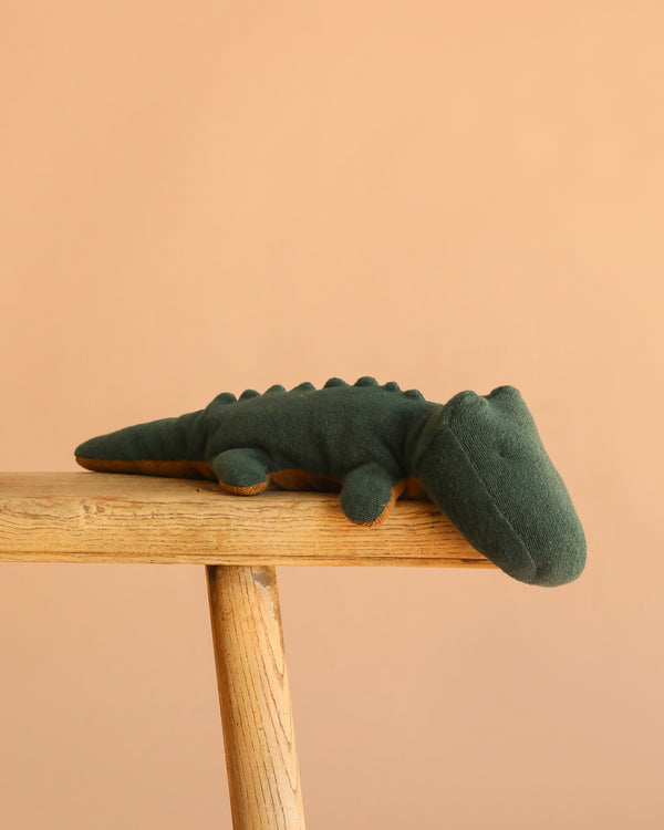 The Maileg Crocodile, Small in Forest Green, a huggable stuffed animal, rests on a wooden bench against a plain beige background.