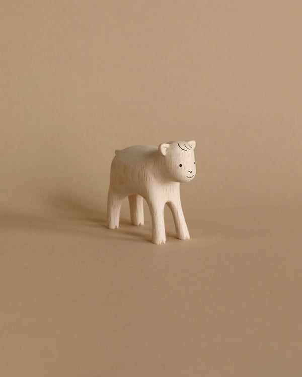 A small, hand-carved wooden figurine of a goat on a plain beige background. The goat, crafted from Albizia wood, has a simplistic design with a smiling face and four legs.
