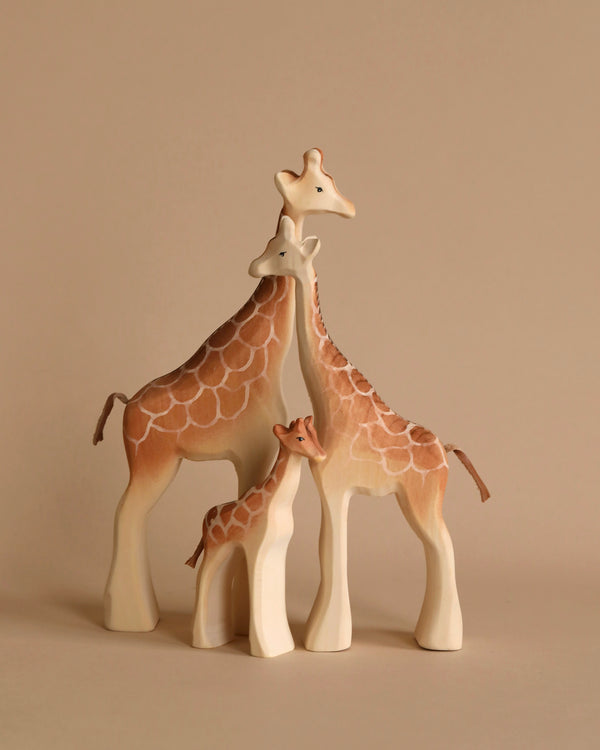 Three wooden giraffe figurines are shown on a neutral background. Two taller giraffes, representing adults, stand protectively on either side of a smaller calf. The Bumbu Wooden Giraffe Family Set features a cream and brown color palette with a simplified, elegant design reminiscent of Montessori aesthetics.