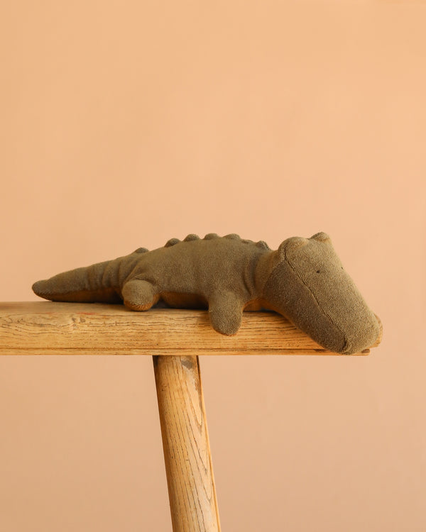 The Maileg Crocodile, Small - Olive Green is a cuddly plush toy that rests on a wooden bench against a beige background, making it an irresistibly huggable choice for fans of soft stuffed animals.