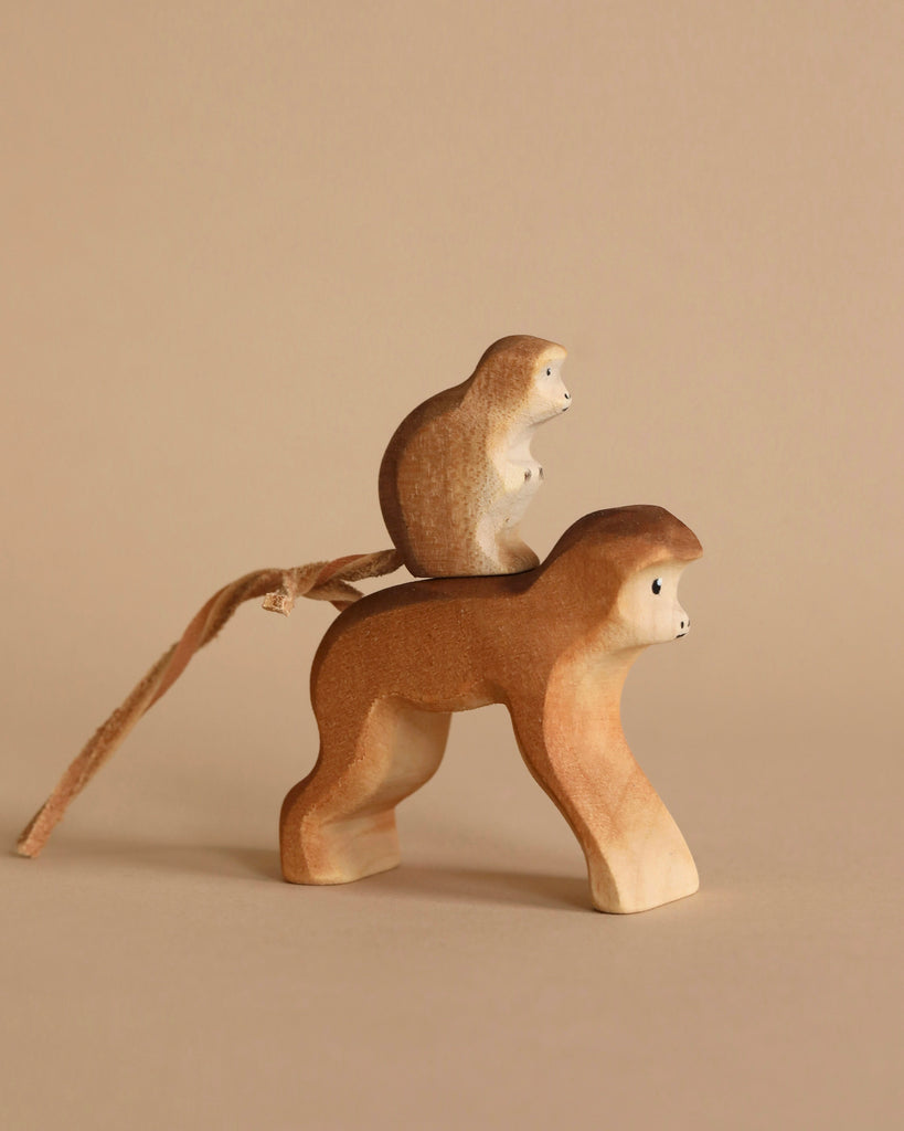 Two wooden toy monkeys against a beige background. The smaller monkey is sitting on the back of the larger monkey. Both figures, part of the Bumbu Wooden Monkey Set, have smooth, rounded features and simple, painted facial details. The larger monkey has a string attached to its body as a tail.