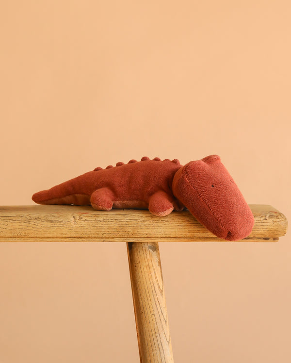 The Maileg Crocodile, Small - Vintage Rose plush in a soft pink hue rests on a wooden bench with a simple peach background.