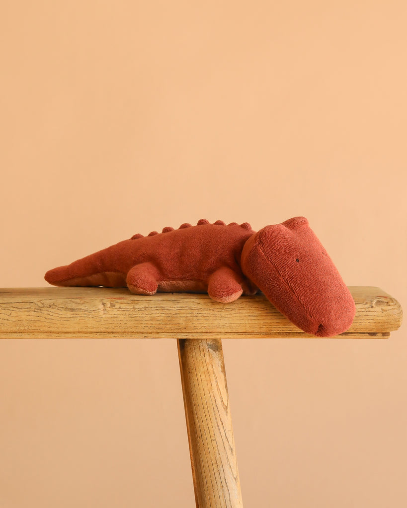 The Maileg Crocodile, Small - Vintage Rose plush in a soft pink hue rests on a wooden bench with a simple peach background.