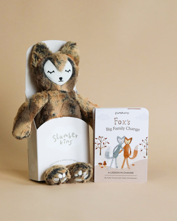 A Slumberkins Fox Kin + Lesson Book On Family Change with hypoallergenic fiberfill next to its packaging and a children's book titled "fox's big family change," all placed against a beige background.